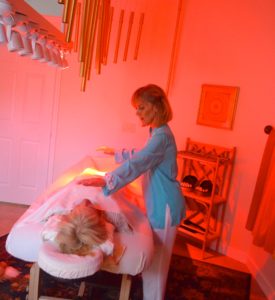 lady in an orange lit up room with wind chimes, healing, meditation room session with a person laying on a massage table.