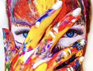 A person with blue eyes with multicolored paint all over her hands and face.